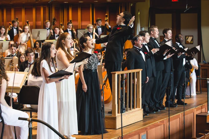 A conductor wearing glasses and formal black attire with a red pocket square stands elevated on a platform with his hand raised, directing a mixed choir. The singers\u2014several women in white and black dresses and men in black suits\u2014stand at microphones watching the conductor with open mouths, singing. An orchestra with strings instruments is visible in the background. The concert hall features warm orange-gold lighting, wooden railings in the foreground, and cream-colored acoustic panels on the walls.