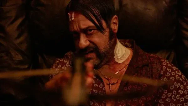 A still from the film Dante. A white man with Italian features, wearing a patterned burgundy dressing gown and a crucifix, aims a wooden crossbow. He has bandages on his head and neck.