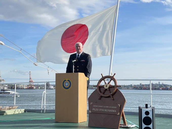 A uniformed person stands behind a podium on a vessel deck, in front of a large Japan flag, with calm water and a city skyline in the background.