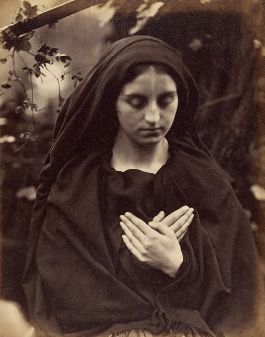 A young woman in dark garb poses with her gaze toward the ground and her hands crossed over her chest, evoking religious imagery.