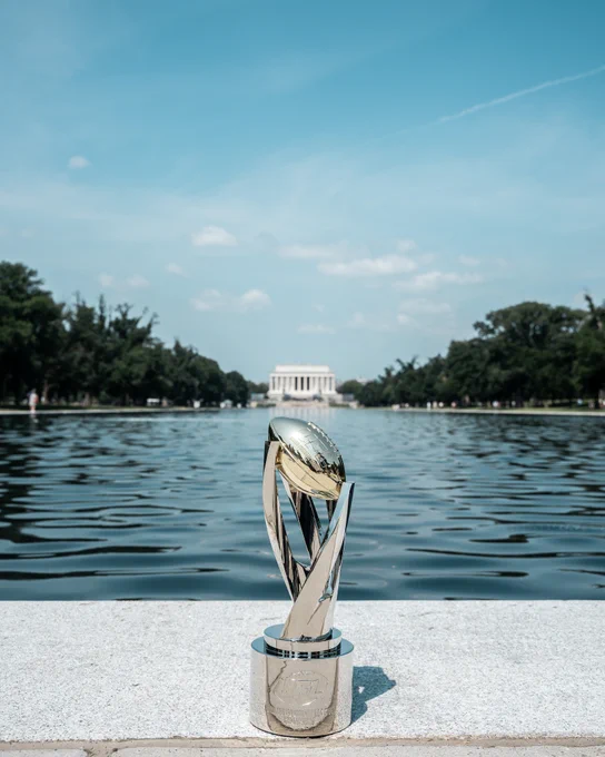 A sleek trophy with a golden top is placed by the water's edge, with a historical monument in the background. The scene conveys a sense of achievement and prestige under a clear blue sky.