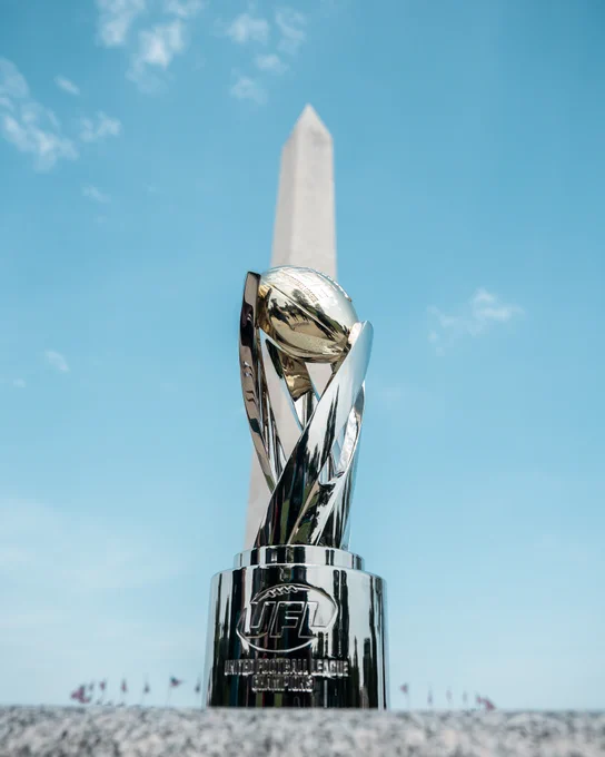 A shiny UFL trophy is displayed against a backdrop of a tall monument under a clear blue sky. The scene conveys a sense of achievement and honor.