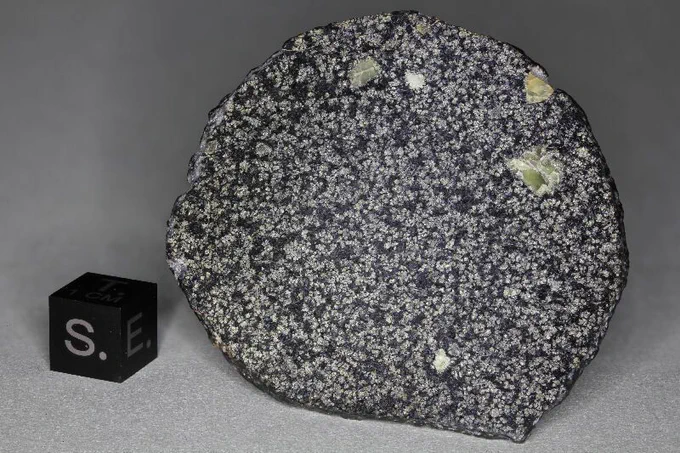 Large slice of a round meteorite revealing white crystals in a black matrix. Several larger green inclusions can be found near the top and the top right. A comparison cube is on the left, showing the orientations of south (out from the sample) and east (to the left) at the time the meteorite was located.