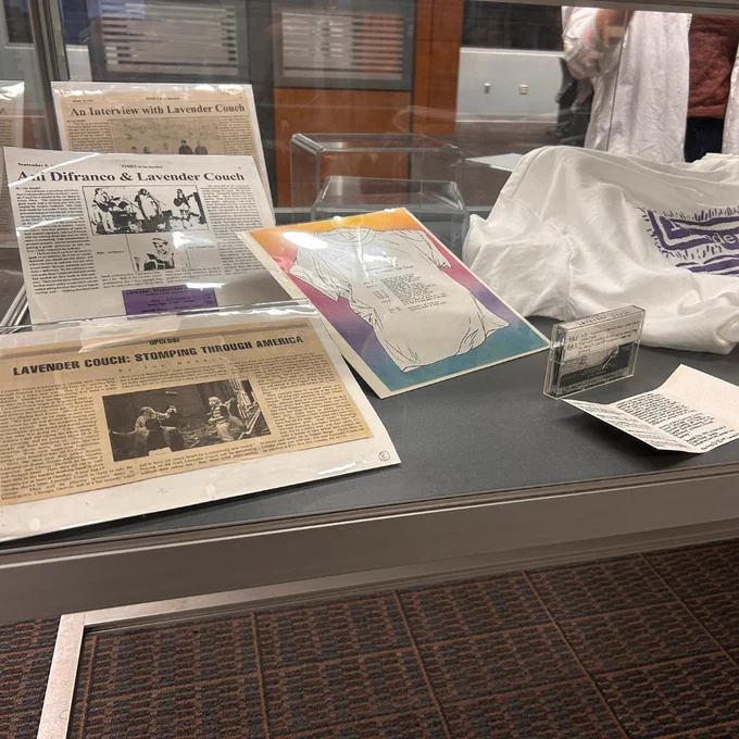 A color photo of a close-up of the Lavender Couch display case. Pictured here are band fliers, news articles, a cassette tape, and a t-shirt. This alt text description was written by an ASC employee without the use of AI. 