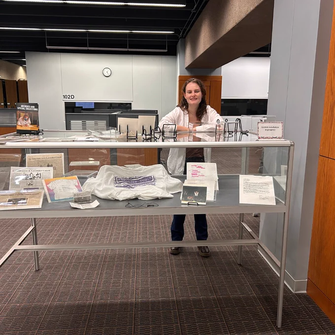 Color photo of ASC intern Britta standing behind a display case filled with fliers, photos, and memorabilia from the band Lavender Couch. Britta is looking at the camera and smiling.  This alt text description was written by an ASC employee without the use of AI. 
