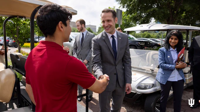 A group of people are outside near golf carts. Two individuals are shaking hands; Undersecretary Nicholas Kent is wearing a suit and the other a red shirt. Others nearby, including Chancellor Latha Ramchand, are engaged in conversation. The Indiana University logo is visible in the bottom corner.