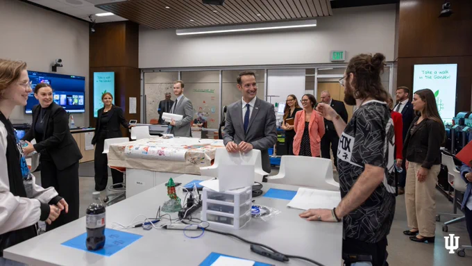 A group of people engaged in lively discussion and interaction in a modern classroom setting. Tables are equipped with various items, including a 3D printer. The atmosphere appears collaborative and educational. The Indiana University logo is visible in the foreground.