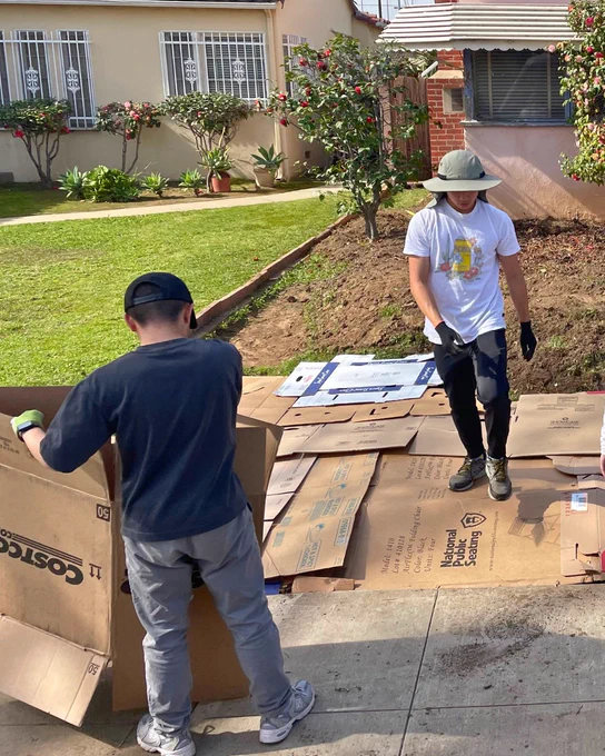 Two individuals are arranging and securing cardboard boxes on a patch of ground near a house with a garden. One person is standing wearing a hat, while the other is bending over a box labeled "Costco Wholesale." The background features a lawn, plants, and a building with bars on the windows.