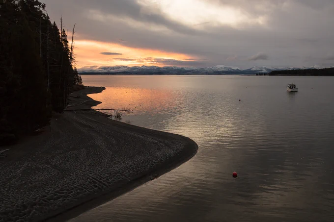 Sunrise above a large lake. Snow-capped mountains can be seen in the distance. A small boat is seen on the water.