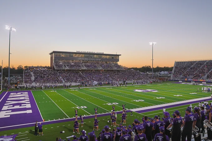A football game at Tarleton Memorial Stadium during sunset. The stands are filled with spectators, and cheerleaders are performing on the sidelines. The field is marked with the name "Texans" in the end zone.