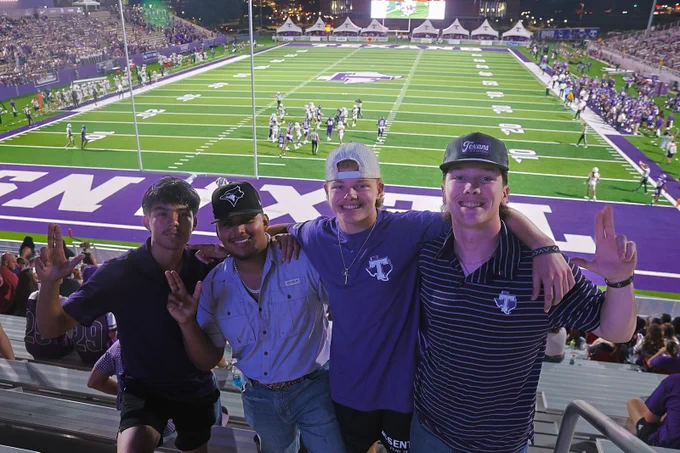 A group of four people stand closely together, smiling in a stadium with a football field in the background. They are wearing casual clothing, some with hats, and are showing hand signs. The field, surrounded by spectators, features the Tarleton State University logo. It's evening, and stadium lights illuminate the scene.