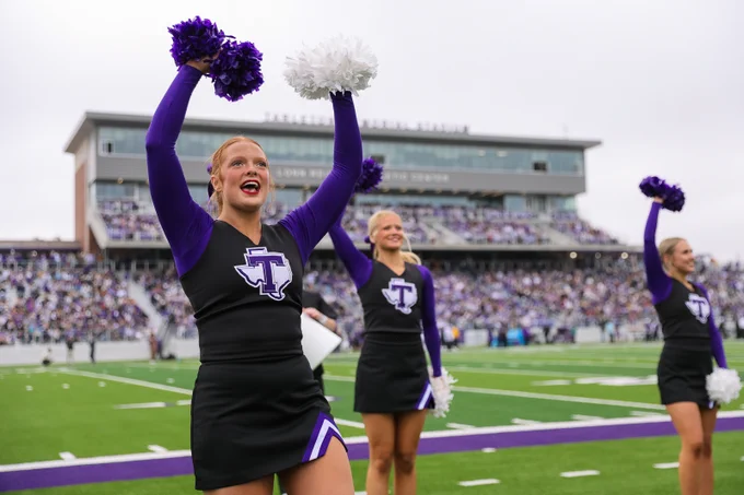 Cheerleaders in black and purple uniforms perform energetically on a football field. The crowd fills the bleachers of the Lonn Reisman Athletic Center in the background.