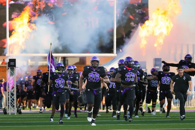 A football team in black and purple uniforms runs onto the field with a player carrying a flag. Flames shoot into the air in the background, creating a dramatic entrance.
