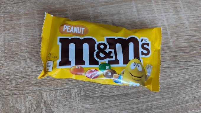 Eine gelbe Packung M&M‘s der Sorte „Peanut“ auf einer eichefarbenen Tischplatte.