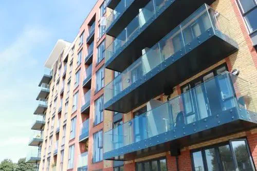 Modern apartment building with brick walls and glass balconies under a clear blue sky. The structure is bathed in sunlight, evoking a sense of urban living.