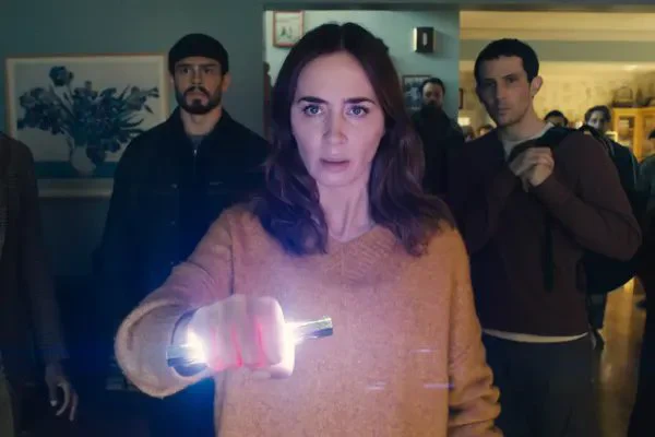 A group of people stand in a room, all looking straight ahead. Foremost is a white woman with long dark hair and an apricot jumper. She is holding out a glowing object in front of her.