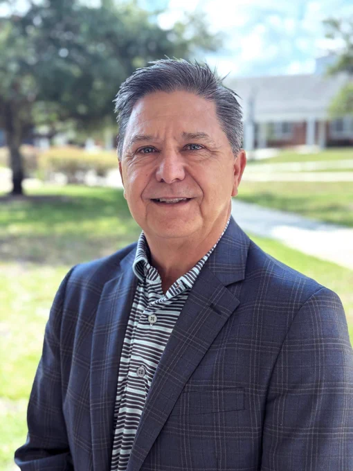 Professional wearing a blazer standing outdoors with a campus blurred in the background.