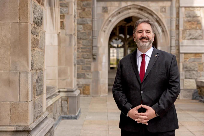 An individual in a suit standing outside a stone building with arched architecture, possibly on an academic campus.