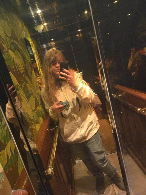 me leaning in a gold-accented elevator, wearing jeans and a white hoodie with a null set on it