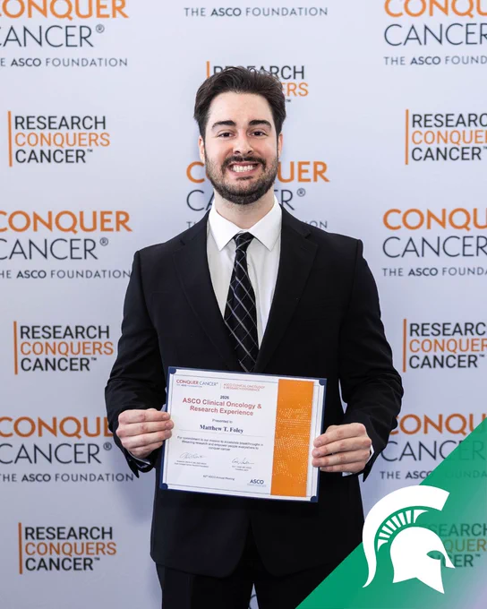A person in a black suit and patterned tie stands in front of a step-and-repeat backdrop printed with “Research Conquers Cancer” and “The ASCO Foundation.” The person holds a certificate that reads “2026 ASCO Clinical Oncology & Research Experience” with the name “Matthew T. Foley” visible. A green graphic with a Spartan helmet logo appears in the bottom right corner of the image.
