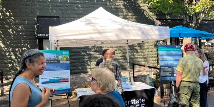 A group of people are gathered around an outdoor booth with a white canopy. The booth has informational posters for the IBR Program, and several people are engaged in conversation, with some looking at the displays. Trees and a building are visible in the background.