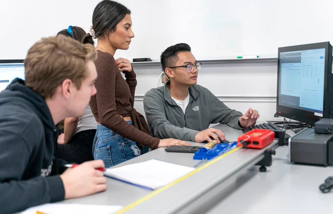 Three people are gathered around a desk in a classroom setting. One is seated at a computer, engaged with a graph on the screen. Two others are attentively watching. A track with small model vehicles is visible in the foreground.