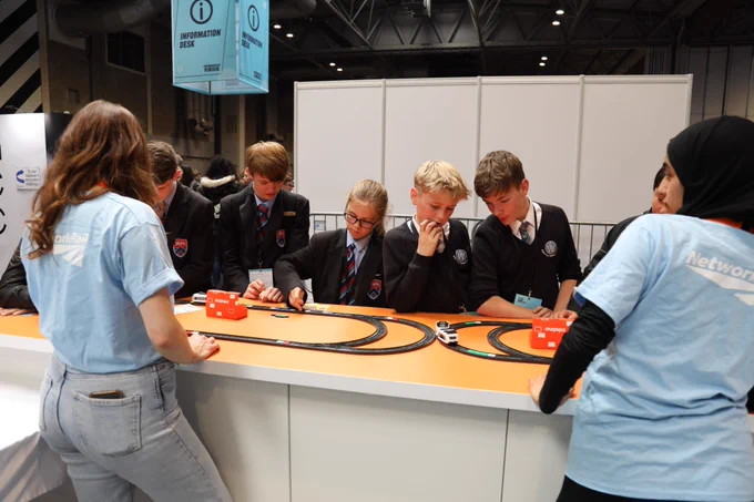 Two colleagues wearing blue Network Rail t-shirts standing at a STEM stall at the Big Bang Fair teaching a group of school pupils about coding.