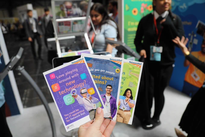 A woman's hand holding three STEM booklets with Big Bang Fair colleagues in the background.