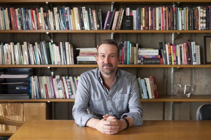 Peter Coviello sitting at a wooden table with hands clasped, in front of a large bookshelf filled with various books in a cozy room.