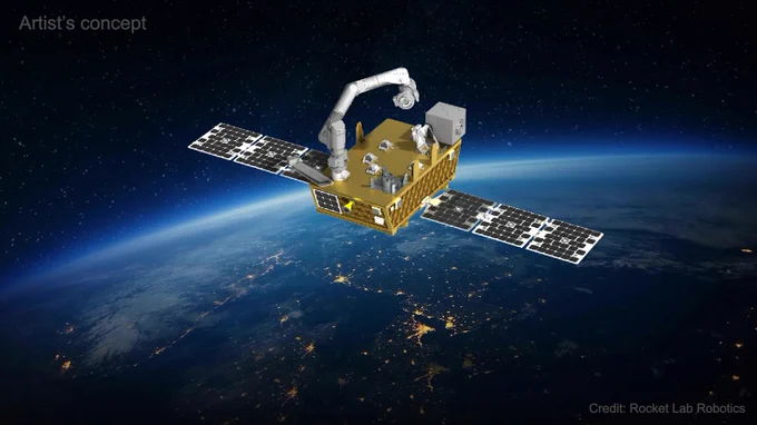 In this illustration of NASA’s Fly Foundational Robots mission, a rectangular golden spacecraft sports a robotic arm extending from the bottom-left corner. The arm is reaching toward a small gray box attached to the spacecraft’s upper-right corner. Two narrow solar arrays extend to either side. The lights of various cities shine on Earth below the satellite, and the limb of the atmosphere glows blue in the distance. The image is watermarked “Artist’s concept” and “Credit: Rocket Lab Robotics.”