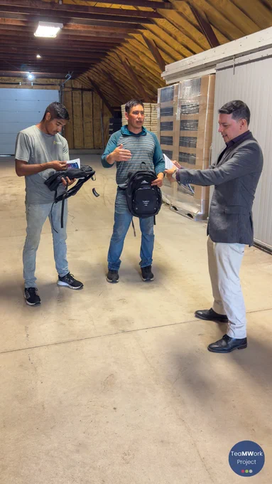 Inside a farm storage building, three individuals stand in a large open space with stacked pallets behind them. One individual distributes TeaMWork Project materials and backpacks while two others review printed information about available support services.
