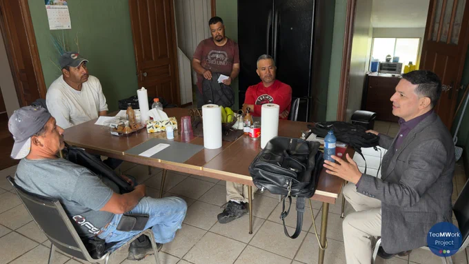 A small group sits around a kitchen table inside a farm residence, with one individual presenting information while others listen. TeaMWork Project materials, food items, and household objects are spread across the table during an informational visit about available worker support services.
