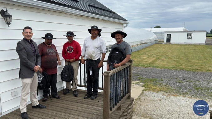 Five individuals stand together on a wooden porch beside a farm building in Chatham-Kent, with greenhouses and fields visible in the background under a cloudy sky. Several individuals hold TeaMWork Project backpacks while gathered during a rainy-day farm visit connecting with seasonal agricultural workers.

