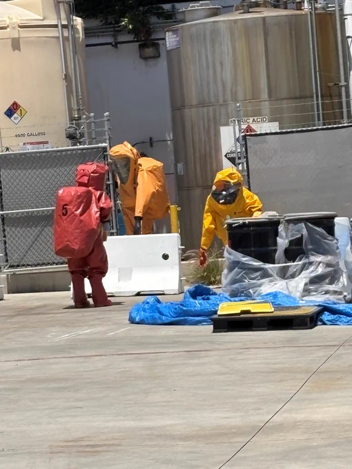 Members of the Huntington Beach Fire Department Hazardous Materials Team conduct a training scenario at an industrial facility while wearing fully encapsulating chemical protective suits. Personnel operate near chemical storage tanks and simulated hazardous materials containers as part of a mitigation exercise.