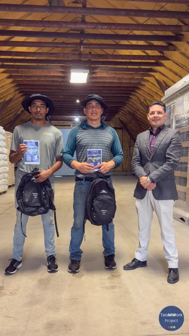 En el interior de un edificio agrícola, tres personas posan de cara a la cámara con folletos y mochilas del Proyecto TeaMWork en las manos. El espacio, con vigas a la vista y materiales apilados, captura un momento de la actividad de divulgación dirigida a los trabajadores agrícolas temporeros.
