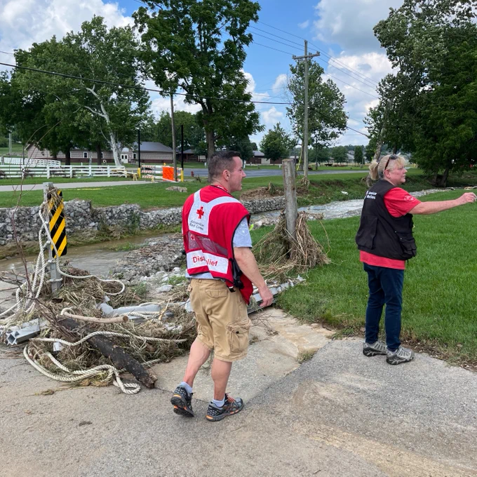 Two Red Cross volunteers assess damage outside on the edge of a grassy area. A stream runs high behind them, and there is debris in the road.