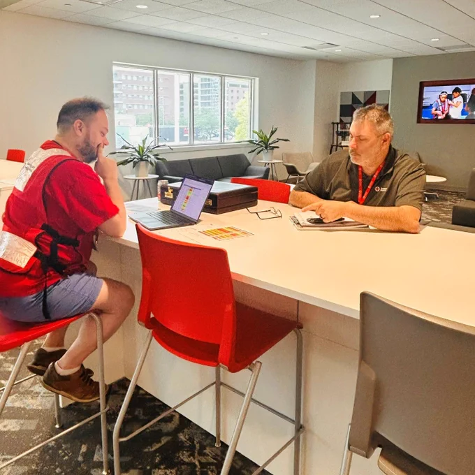 Two Red Cross volunteers sit at a table indoors, with laptops and paperwork in front of them.