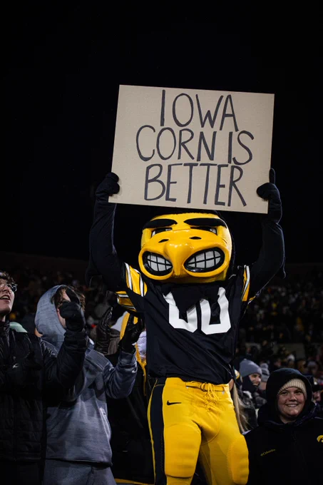 Iowa Hawkeye mascot holding a sign that says 'Iowa Corn is Better' at a nighttime sports event.