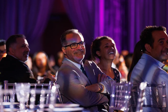 A group of people seated at a formal event with purple lighting. They are smiling and listening to  a presentation. 