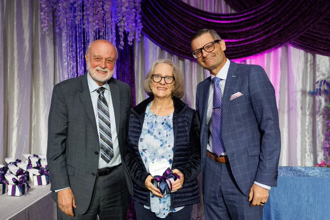 Three LLUH employees are posing together and smiling in a formal setting with elegant purple and silver drapery. They are dressed in business attire, and the person in the center is holding a small, wrapped gift.