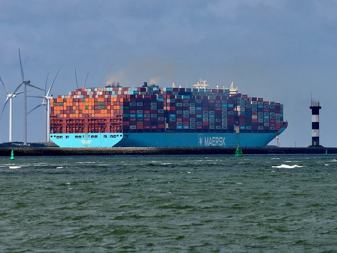 A massive container ship loaded with colorful containers sails past wind turbines and a lighthouse. The scene conveys a sense of maritime industry and global commerce.