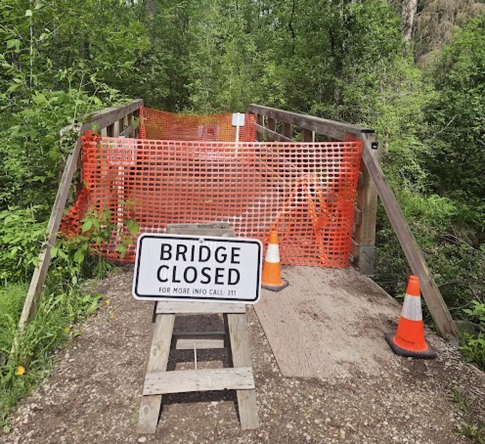A pedestrian footbridge in a wooded area is blocked by orange safety fencing and a wooden A-frame sign that reads BRIDGE CLOSED FOR MORE INFO CALL: 311