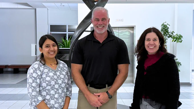 Three colleagues smile for a photo in a bright modern building lobby featuring a large double-helix sculpture in the background. From left to right: a woman in a gray floral blouse, a tall man in a black polo shirt, and a woman in a black jacket and red top.