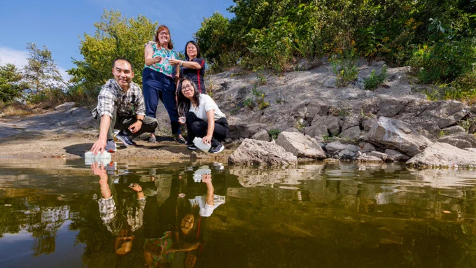 Four researchers pose at the edge of a shallow body of water outdoors, with their reflections visible on the water's surface. Two crouch near the water holding sample collection bottles while two others stand behind them. Rocky terrain and green vegetation are visible in the background under a blue sky.
