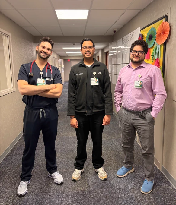 Three medical professionals stand in a hospital hallway. Two are in medical attire, one in a pink shirt. They wear ID badges and are smiling. Brightly colored decorations hang on a board in the background.