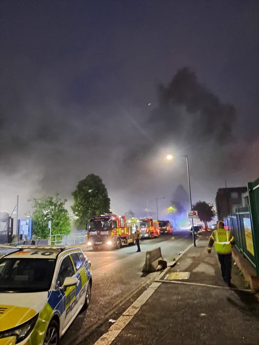 Photo of three fire engines parked on a street in the evening, with a police care in the foreground. Heavy smoke is visible in the background.