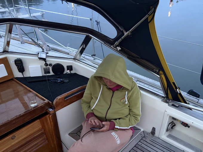 Ein Mädchen in Jogginghose (pink) und Hoodie (grün) mit Kaputze über dem Kopf sitzt im Cockpit eines Segelbootes und tippt auf ihrem Handy. Man sieht Teile des Bootes (etwas Holz des Niedergangs und die Sprayhood). Wichtig: Es ist taghell!