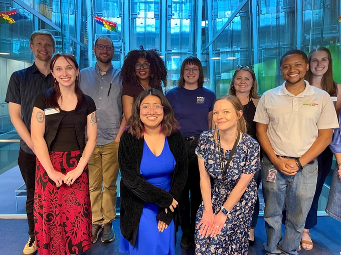 A group of ten diverse AmeriCorps members stand together smiling inside a modern glass building atrium with blue glass paneling.