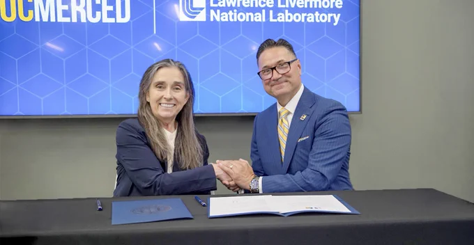 Two professionals shaking hands over a signed agreement at a table with UC Merced and Lawrence Livermore National Laboratory logos in the background.