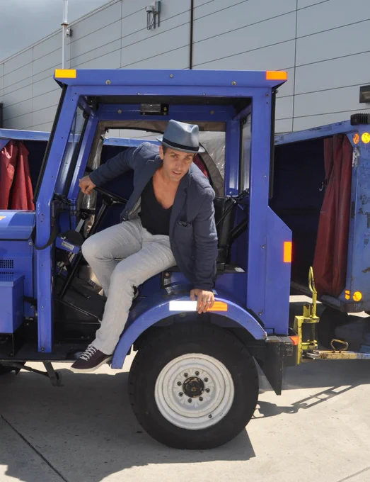 A person wearing a gray hat and jacket is stepping out of a blue airport luggage tug. The background shows a gray building.